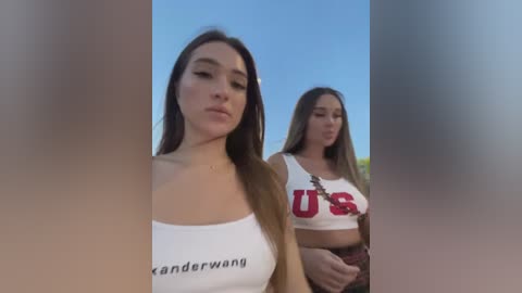 Media: Video of two young women in white crop tops with red \"US\" lettering. One has long brown hair, the other has long blonde hair. They are outdoors against a clear blue sky.