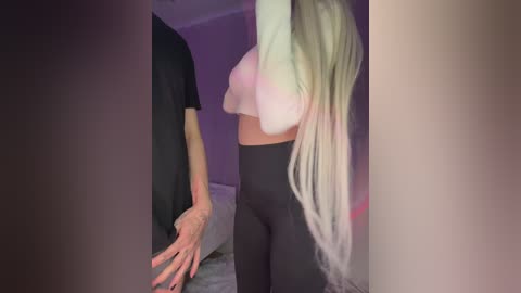 Media: Video of a slender, light-skinned woman with long, platinum blonde hair, wearing a white crop top and tight black leggings, standing in a dimly lit room with purple walls.