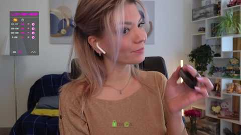 Media: A video of a young woman with light skin, long blonde hair in pigtails, wearing a beige top, holding a phone, in a cozy living room with a TV, shelves, and plants.