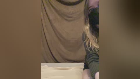 A video of a person in a beige tank top and dark green sweater, with long blonde hair, bending over a white surface, possibly a table or desk.