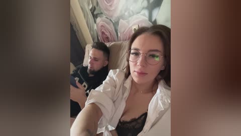 Media: A video shows a Caucasian woman with brown hair and glasses, wearing a white blouse, taking a selfie with a man in a black shirt in the background.
