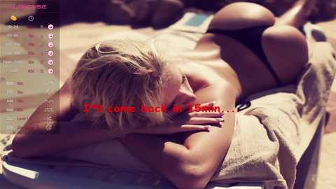 Video of a topless blonde woman lying on a beige towel on a sunlit beach, wearing only black bikini bottoms. Text overlay reads, \"4:30 come back in 5 minutes.\