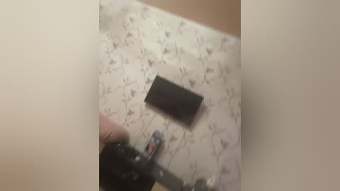 A video shows a hand holding a black box cutter blade against a beige wall with a floral pattern. The hand is partially obscured, and the blade is being applied with pressure, creating a sharp, clean cut.