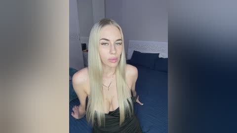 Video of a fair-skinned, blonde woman with long hair, wearing a black, strapless dress, sitting on a blue bed with white headboard, in a dimly lit bedroom.