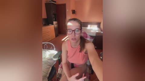 Media: Video of a young woman with glasses and a light complexion, wearing a pink tank top and shorts, sitting on a bed in a cozy, warmly lit bedroom with a wooden dresser and a partially visible TV.