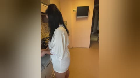 Media: Video of a woman in a white bathrobe, with long black hair, standing at a stove in a small, beige-walled kitchen. A TV is mounted on the wall above the stove.