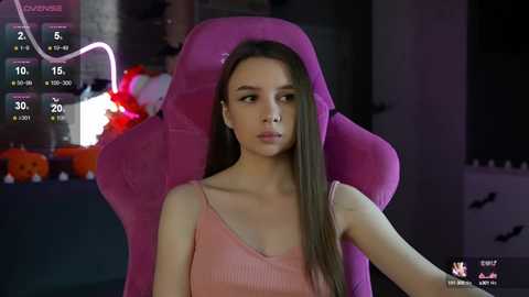 Media: Video of a young woman with long brown hair wearing a pink tank top, seated in a pink gaming chair, with Halloween decorations and a digital scoreboard in the background.