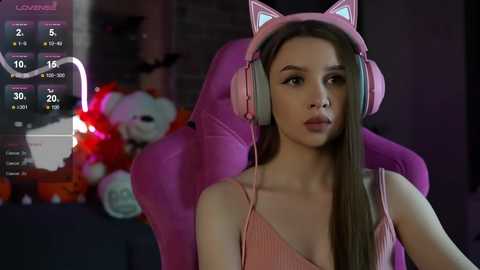 Media: Video of a young woman with long brown hair, wearing pink headphones with cat ears, sitting in a pink gaming chair. Background shows a dimly lit room with a stuffed animal and a clock displaying 5:18.