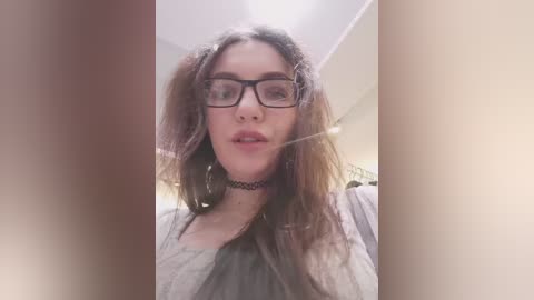 A close-up video of a young woman with long, tousled brown hair, wearing black-framed glasses, a black choker, and a light gray shirt. Background is blurry and out of focus.