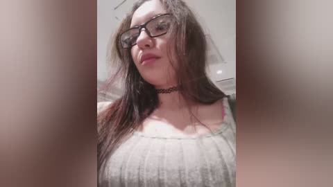 A video of a fair-skinned woman with long brown hair, wearing black-framed glasses and a black choker necklace, in a gray ribbed top. The background is blurred.