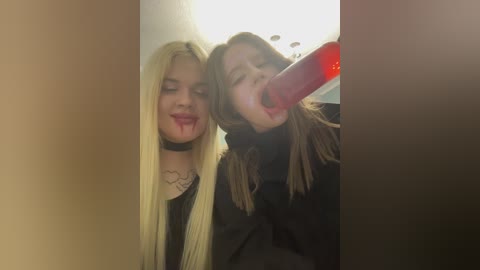 Video of two young women, one with long blonde hair and the other with brown hair, wearing black outfits, both licking a large red dildo in a dimly lit room.