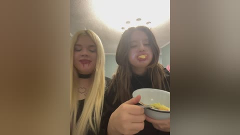 Video of two young women with long blonde and brunette hair, respectively, licking ice cream cones in a dimly lit room with a white ceiling light.