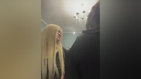Video of a woman with long blonde hair and a black jacket, standing in a dimly lit hallway with a chandelier overhead, next to a man in a dark coat.
