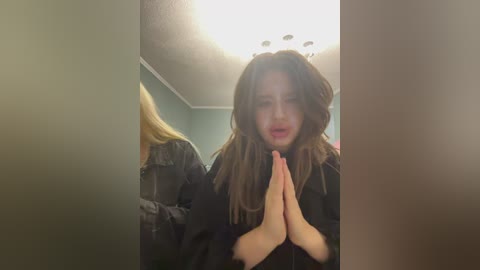A video of a young woman with long, wavy brown hair, wearing a black jacket, praying with clasped hands, standing in a dimly lit room with a ceiling light.