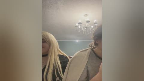 Video of a young woman with long blonde hair, wearing a black choker, standing in a dimly lit room with a chandelier, holding a beige towel.
