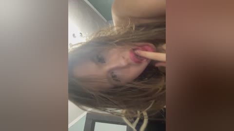 Video of a young woman with messy, light brown hair and pink lipstick, holding a wooden stick near her mouth, partially obscured by her hair. The background shows a green wall and a white ceiling.