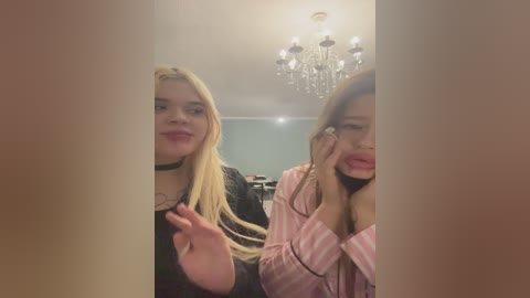Video of two young women with long blonde and brunette hair, smiling, one in black, the other in pink striped pajamas, in a dimly lit room with a chandelier.