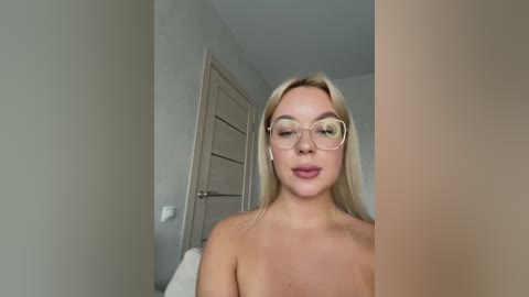 Media: Video of a blonde woman with fair skin, wearing large, round glasses, topless, in a room with beige walls and a white door.