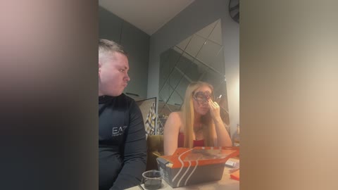Media: A video showing a blonde woman with glasses and a man with short hair at a table in a dimly lit room. They are both wearing dark clothing.
