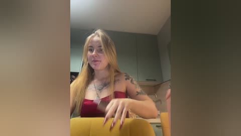 Media: Video of a blonde woman with fair skin, wearing a red strapless top, sitting in a modern kitchen. She has tattoos on her arms and a serene expression.