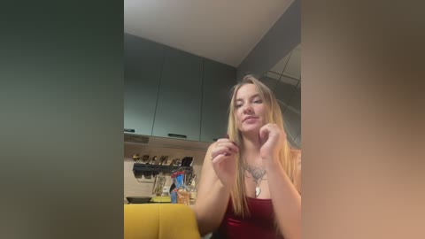 Media: Video of a blonde woman with long hair, wearing a red top, smiling, with a tattoo on her chest, standing in a modern kitchen with grey cabinets and a yellow table.