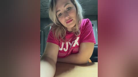 A video of a blonde woman with fair skin, wearing a red T-shirt, leaning forward on a wooden table. The background shows a dimly lit room with grey walls.