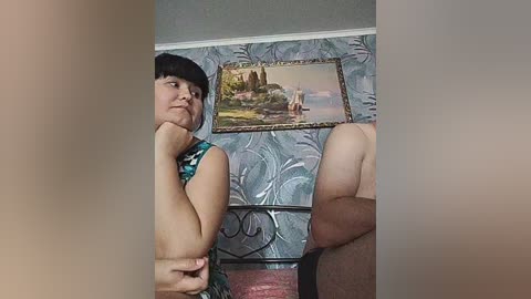 A video of a smiling Asian woman with short black hair, wearing a colorful dress, standing next to a man with a muscular build. Background features a decorative wall with a framed landscape painting.
