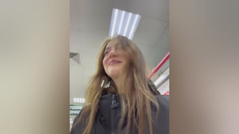 Media: Video of a young woman with long, light brown hair, smiling with eyes closed, wearing a black jacket, standing in a well-lit, modern indoor setting with fluorescent lighting and visible ceiling panels.