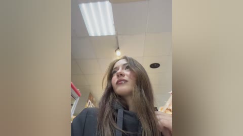 Media: Video of a woman with long brown hair in a black robe, standing in a salon with white tiled walls, fluorescent lights, and a red chair in the background.