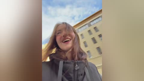 Media: Video of a smiling woman with light brown hair, wearing a dark coat, standing outdoors in front of a yellow building with multiple windows under a clear blue sky.