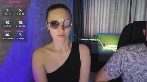 A video of a fair-skinned woman with a slim build, wearing a black one-shoulder dress, and large, round sunglasses, sitting in a dimly lit room with a green screen and a person partially visible in the background.