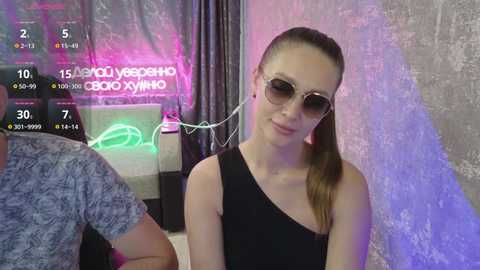 Video of a woman with fair skin, long brown hair in a ponytail, wearing sunglasses, black one-shoulder top, and headphones, sitting in a dimly lit room with a colorful neon sign.