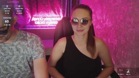 A video of a woman with long brown hair in a ponytail, wearing sunglasses, a black one-shoulder dress, seated in a dimly lit room with neon pink lights and a \"Porsche Experience Center\" sign.