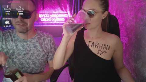 A video of a man in a grey shirt and woman in a black dress, both holding drinks, in a dimly lit bar with neon lights and a TV screen displaying game scores.