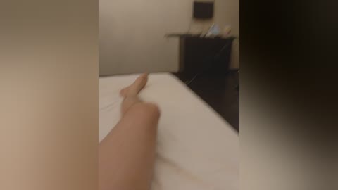 A video captures a close-up view of a person's bare legs and feet on a white bed, with blurred background details of a bedroom, including a dresser and a wall-mounted TV.