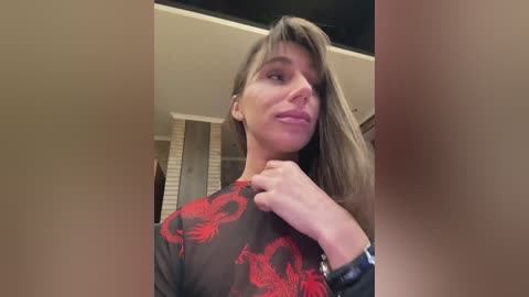 Video of a light-skinned woman with long, straight brown hair, wearing a sheer black top with red dragon print. She is indoors, standing with her left hand on her chest, in a dimly lit room with beige walls and striped blinds.