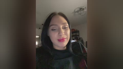 A video of a young woman with fair skin and long black hair, wearing dark clothing, winks with red lipstick, set against a dimly lit room with shelves in the background.