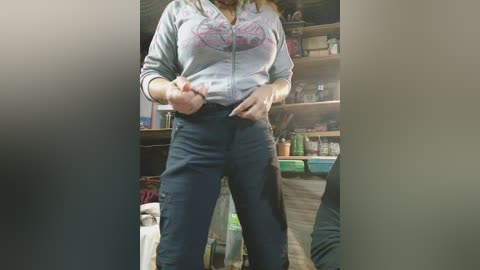 Media: Video of a woman in a gray hoodie, dark pants, and white sneakers, standing in a cluttered storage room with shelves full of boxes and containers.