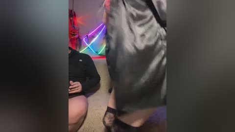 Video of a woman in a black jacket, kneeling on a carpet, with a colorful neon sign and a jack-o'-lantern in the background.