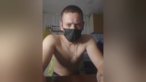 Media: Video of a shirtless man with short brown hair, wearing a black mask and green pants, leaning forward, in a dimly lit room with cabinets and a yellow towel hanging on the wall.