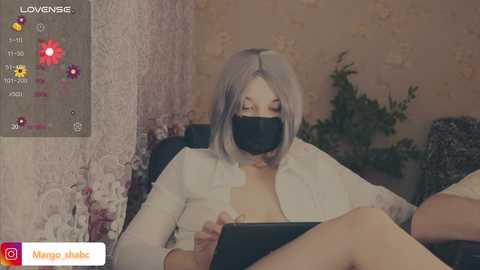 Video of a woman with shoulder-length platinum blonde hair, wearing a white blouse and black face mask, seated on a black chair in a room with floral wallpaper.