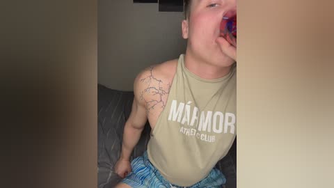 Media: Video of a young man with a tattoo on his left shoulder, wearing a \"MAI MORI\" tank top and blue shorts, eating a blue popsicle, seated on a gray bed.