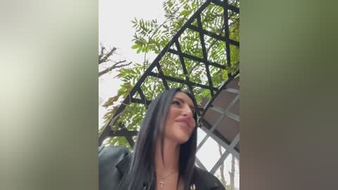 Media: Video of a young woman with long dark hair, wearing a black jacket, smiling, standing in a lush garden with a metal lattice structure in the background.