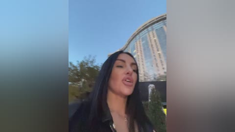 Media: A video of a woman with long, straight, dark hair, wearing a black leather jacket and a black top, with a clear blue sky and a large, modern building in the background.