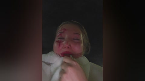 A dimly lit video of a blonde woman with blood on her face, clutching a white cloth. She appears distressed, with her eyes half-closed.