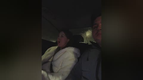 A dimly lit video captures a terrified woman with short dark hair and a white jacket, hugging a man with short dark hair in a car. The background is dark, with blurred details.