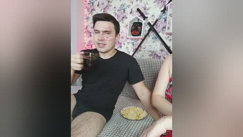 Video of a young man with a bloody face, wearing a black t-shirt and shorts, drinking a glass of cola, sitting on a floral-patterned couch, surrounded by a red-lit room.