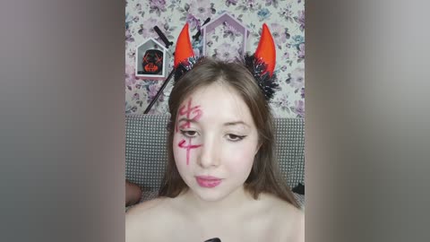 Video of a young woman with light skin and straight brown hair, wearing an orange devil hat with black horns and a pink face mask with Japanese kanji. She has an expressionless face.