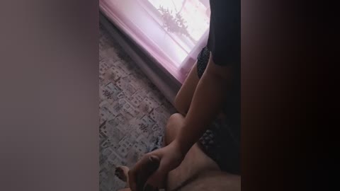 A video of a dimly lit bedroom with a man wearing black shorts and a woman in a black bra, sitting on a patterned carpet. The room has a small window with sheer pink curtains.
