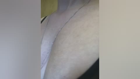 A close-up video of a light-skinned person's upper torso, showing a thin silver necklace and a hint of a white bra strap. The background is blurred, with a pale, neutral tone.